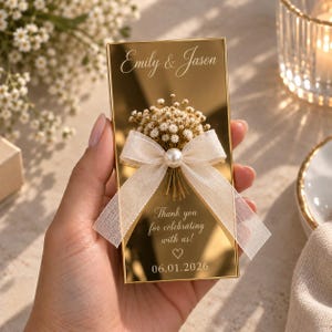 May include: A rectangular gold mirror favor featuring the engraved text Emily & Jason, Thank you for celebrating with us!, and 06.01.2026. A small bouquet of dried white flowers is attached to the center, accented by a sheer white ribbon and a pearl.