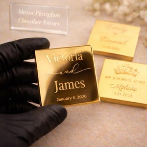 Könnte beinhalten: Goldfarbene quadratische Gastgeschenke mit eingravierten Namen und Daten. Eines trägt die Aufschrift "Victoria and James, 6. Januar 2026". Ein anderes trägt die Aufschrift "Emily und Daniel, 10.03.2023". Ein drittes trägt die Aufschrift "Melissa, 2.03.2023."