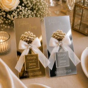 May include: Two rectangular mirrored signs, one gold and one silver, each featuring a white ribbon bow, a small bouquet of dried flowers, and a square plaque. The text reads Victoria and James with the date January 6, 2026, and 06/06/2026.