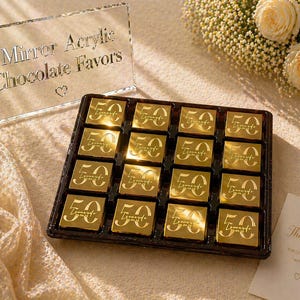 May include: A tray of gold-wrapped chocolates, each marked with "50", sits beside a clear acrylic sign that reads "Mirror Acrylic Chocolate Favors". A small card says "Thank You". The chocolates are a celebratory treat.