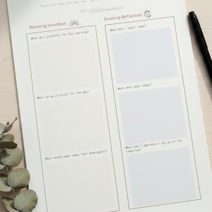 May include: A beige Daily Meditation journal page with sections for Morning Intention and Evening Reflection. The page includes prompts such as "What am I grateful for this morning?" and "What did I learn today?" A black pen rests on the page.