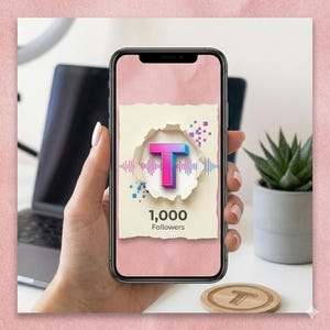 May include: A smartphone displays a graphic with a large, gradient pink and blue letter "T" emerging from a torn paper effect. The screen reads "1,000 Followers" with sound wave graphics. A wooden coaster with a "T" is also visible.