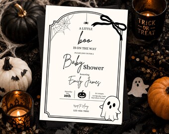 May include: A rectangular white baby shower invitation card with black text and graphics. The card features a spider web, a spider, a bow, a pumpkin, and a ghost. The text reads: A LITTLE boo IS ON THE WAY, Baby Shower, Emily James, Oct 25th.