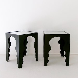 May include: A pair of square side tables featuring a matte black finish. Each table has a flat top surface and four legs with a distinct wavy, scalloped edge design on all sides, creating an arched opening between the legs of the furniture.
