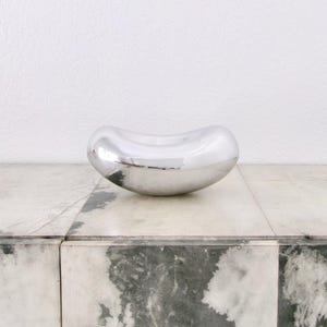Op de afbeelding: Een gepolijste, zilverkleurige metalen kom met een gladde, gebogen, boonachtige vorm. Het oppervlak is sterk reflecterend en creëert een spiegelende afwerking die het omgevingslicht en de omgeving opvangt in zijn ronde, organische vorm.