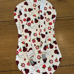 May include: White fabric baby bib with red ladybugs and roses, and attached suspenders with silver metal clips. The bib is patterned with ladybugs, red roses, and green leaves.