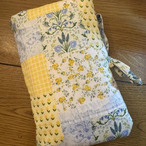 May include: A rectangular fabric pouch featuring a patchwork design with various patterns in yellow, blue, and white. The textile displays floral motifs, small yellow tulips, and a yellow gingham check pattern, finished with a fabric tie closure.
