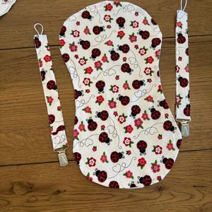 May include: White baby bib and two pacifier clips with a ladybug and floral pattern. The bib is a curved shape with a ladybug and flower print. The pacifier clips have the same pattern and metal clips.