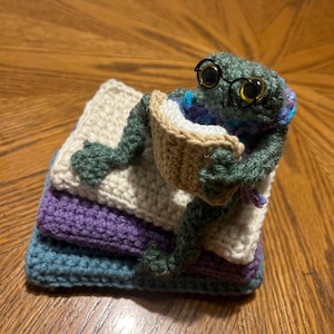 May include: A handmade crochet frog wearing glasses, holding a book, and sitting on a stack of colorful crochet squares. The frog is green, with blue and purple accents. The squares are blue, purple, and cream-colored.