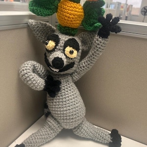 May include: A crocheted lemur plush toy with gray fur, black hands, feet, and facial markings, and yellow eyes. The lemur is holding a crocheted yellow and orange fruit-like object with green leaves above its head with one raised arm.