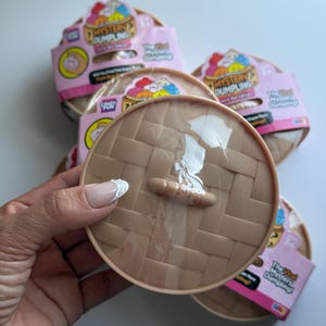 May include: A round, tan plastic container with a woven basket texture and a small handle on top, resembling a bamboo steamer. The packaging features the text Mystery Dumpling and The Viral Mystery Dumpling, with pink accents and colorful graphics.