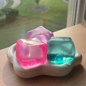 May include: Three translucent, squishy, cube-shaped sensory toys in pink, purple, and teal colors. The teal cube features the embossed text "NEE DOH" on its front face. These items are arranged on a white, flower-shaped, rounded tray.