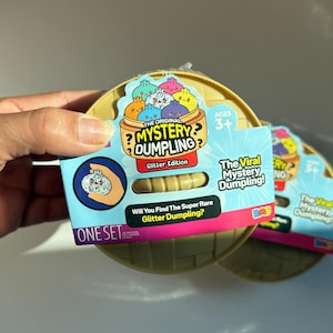 Könnte beinhalten: Das Spielzeugset The Original Mystery Dumpling Glitter Edition in einem runden beigen Behälter. Die Verpackung zeigt bunte Cartoon-Teigtaschen, den Text The Viral Mystery Dumpling und die Frage, ob man das seltene Glitzer-Exemplar findet.