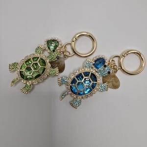 May include: Two bejeweled turtle keychains. One is green, the other is blue. Both have gold-colored metal accents, pearl-like beads, and small seashells. The keychains are designed with a gold-colored clasp.