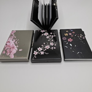 Può includere: Tre porta carte in metallo con disegni floreali. Uno è aperto, mostrando le carte. I porta carte sono grigi, neri e neri, ognuno con un motivo floreale diverso in rosa, bianco e giallo. Il porta carte aperto è nero.