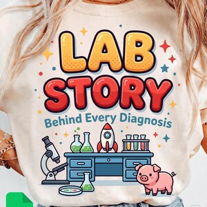 May include: A light beige t-shirt with a colorful graphic that reads "LAB STORY" above "Behind Every Diagnosis." The design includes a microscope, beakers, a rocket, and a cartoon pig. The shirt has a crew neck.