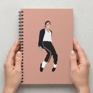 May include: A spiral-bound notebook with a dusty rose cover featuring a minimalist graphic illustration of a person in a black jacket, white shirt, and black trousers with a side stripe, captured in a dynamic dance pose with white socks and black shoes.