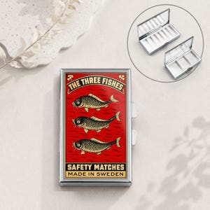 Puede incluir: Un estuche metálico rectangular con un diseño gráfico rojo que incluye tres ilustraciones de peces y el texto THE THREE FISHES SAFETY MATCHES MADE IN SWEDEN. El estuche tiene un marco plateado e incluye una vista interior con dos compartimentos.