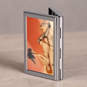 Puede incluir: Una pitillera rectangular de metal con acabado plateado pulido. El panel frontal presenta una ilustración vertical de una figura reclinada con cabello rojo, que lleva medias y tacones negros, interactuando con un gato negro sobre un fondo naranja.