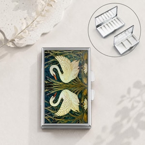 May include: A rectangular metal case featuring a decorative print of two white swans swimming among green reeds and water lilies. The case has a silver-toned frame and a hinged lid. An inset shows the interior with multiple white dividers.