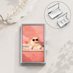 Puede incluir: Un estuche rectangular de metal plateado con el gráfico de un gato esponjoso con gafas de sol naranjas, descansando en una piscina frente a una pared de azulejos rosas con una copa de cóctel. El estuche se abre para mostrar compartimentos.