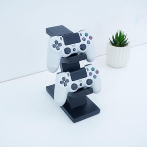 Könnte beinhalten: Ein schwarzer Holzständer für zwei weiße Gamecontroller. Die Controller haben schwarze Knöpfe und farbige Aktionstasten. Im Hintergrund befindet sich eine kleine Topf-Sukkulente.