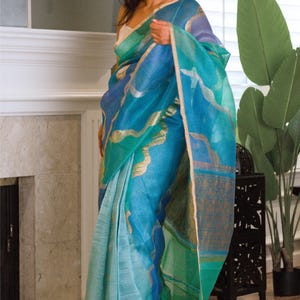 May include: A woman wearing a vibrant blue saree with gold accents. The saree features a gradient of blues and greens, with a shimmering, translucent fabric. The garment is draped elegantly, showcasing its flowing design and intricate details.