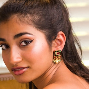 May include: Gold-toned square earrings with red and green accents, featuring a pearl drop. The earrings have a geometric design and are worn on the ear. The jewelry is a statement piece.