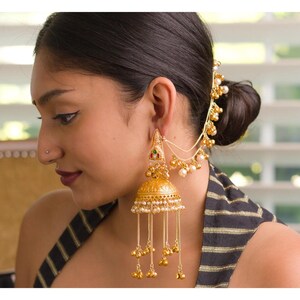 May include: Gold-toned, ornate earrings with dangling beads and a matching headpiece. The earrings feature a bell-shaped design with intricate detailing and small pearl accents. The headpiece connects to the earrings with delicate chains.