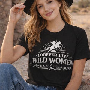 May include: Black t-shirt with the text "FOREVER LIVE WILD WOMEN" and a graphic of a woman riding a horse. The shirt is paired with blue jeans and a black hat.