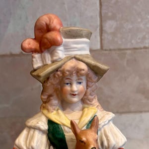 May include: A porcelain figurine of a woman in a long dress holding a fawn. The figurine has a hat with a floral accent, and the woman's dress has green and red accents. The fawn is brown.