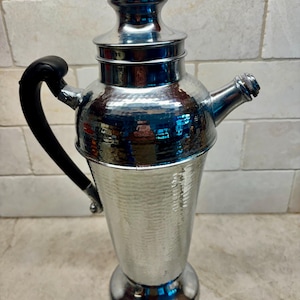 May include: A silver-toned metal cocktail shaker featuring a hammered texture on the body, a smooth domed lid, a tapered base, and a curved black handle attached to the side. The shaker includes a short spout for pouring liquids.