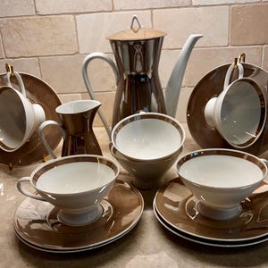 May include: A coffee service set featuring a tall coffee pot with a lid, a creamer, and four cups with matching saucers. Each piece is crafted from white ceramic with a glossy brown band and gold trim accents around the rims and handles.