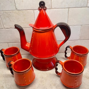 May include: A bright orange ceramic tea set consisting of a tall teapot with a pointed lid and four matching mugs. Each piece features a glossy finish with dark brown gradient accents on the handles, spout, and lid, and a vertical row of raised dots.