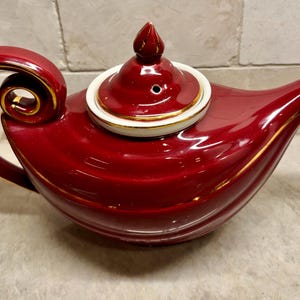 May include: A deep red ceramic teapot featuring a curved handle with a gold-trimmed edge, a matching lid with a pointed finial, and a gold-rimmed spout. The body of the teapot has horizontal ridges and a glossy finish with gold accents.