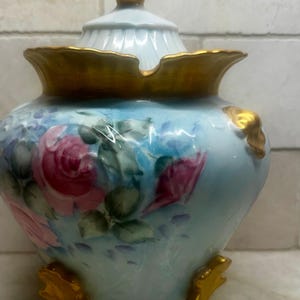 May include: A light blue ceramic jar with a lid, featuring hand-painted pink roses and green leaves. The jar has gold accents on the rim, lid finial, and feet. A decorative piece for home decor.