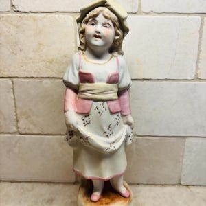 May include: A porcelain figurine of a child wearing a bonnet, a white dress with a floral pattern, and a pink vest. The figure stands on a circular base, featuring painted details in soft pastel colors including pink, yellow, and gold accents.
