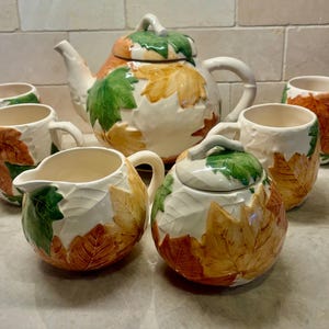 May include: A ceramic tea set featuring a teapot, a creamer, a sugar bowl with a lid, and four mugs. Each piece is cream-colored with embossed, hand-painted autumn leaves in shades of green, yellow, orange, and brown, creating a textured surface.