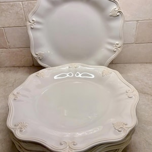 May include: A stack of white ceramic dinner plates featuring a scalloped edge and embossed scrollwork details along the rim. The plates have a glossy finish and a smooth, circular center, with one plate standing upright behind the stacked set.