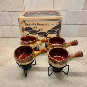 May include: A set of four ceramic butter and sauce warmers with brown and cream glazed finishes, each featuring a small handle and a pouring spout. The warmers sit on black metal wire stands, accompanied by a cardboard box with product text.