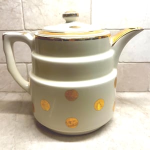 May include: Cream-colored ceramic teapot with a lid and handle, adorned with gold trim and gold polka dots. The teapot has a classic design with a spout for pouring. A vintage-style kitchenware item.