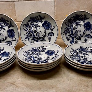 May include: A set of white ceramic saucers featuring a blue onion pattern with floral and botanical motifs. The collection includes three upright saucers and three stacks of saucers, each with a scalloped edge and a central circular indentation.