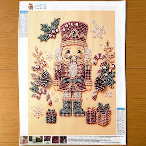 May include: A diamond painting kit on a rectangular canvas featuring a central nutcracker figure surrounded by holly leaves, snowflakes, candy canes, pinecones, and gift boxes. The design includes a color legend and the text ZZH163 Size 30*40.