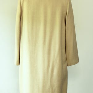 Puede incluir: Un abrigo beige claro, hasta la rodilla, con un sutil patr&oacute;n texturizado. El abrigo presenta un cuello cl&aacute;sico, mangas largas y un corte entallado. La prenda se exhibe en un maniqu&iacute; vintage.