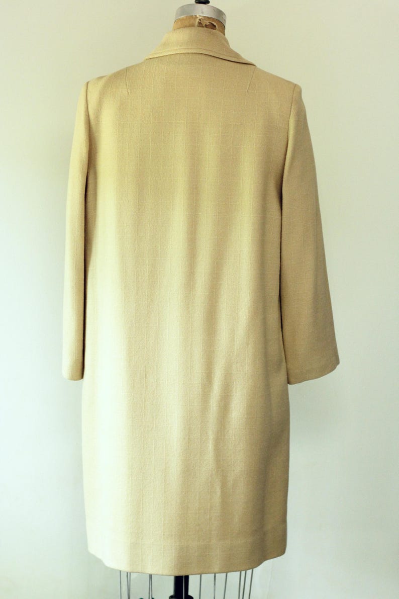 Puede incluir: Un abrigo beige claro, hasta la rodilla, con un sutil patr&oacute;n texturizado. El abrigo presenta un cuello cl&aacute;sico, mangas largas y un corte entallado. La prenda se exhibe en un maniqu&iacute; vintage.