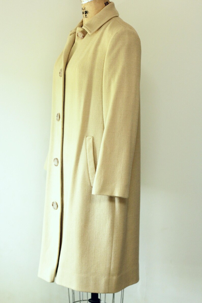 Puede incluir: Un abrigo largo de color beige claro con cuello con solapas y tres botones. El abrigo tiene un solo bolsillo y se exhibe en un maniqu&iacute;. El abrigo est&aacute; hecho de una tela texturizada.