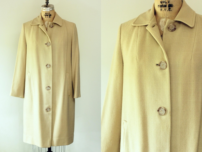 Puede incluir: Un abrigo beige vintage con un dise&ntilde;o cl&aacute;sico. El abrigo presenta un cuello, mangas largas y una botonadura frontal. Tiene un corte entallado y dos bolsillos delanteros. El abrigo se exhibe en un maniqu&iacute;.
