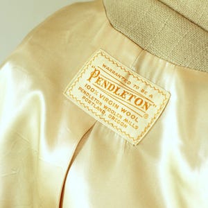 Puede incluir: Primer plano del forro de sat&eacute;n color crema de una chaqueta, con cuello beige. Una etiqueta rectangular dice "PENDLETON" y "100% Virgin Wool, Pendleton Woolen Mills, Portland, Oregon."