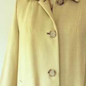 Puede incluir: Primer plano de un abrigo beige claro con cuello con solapas y tres botones grandes y redondos. El abrigo tiene una sutil textura de rayas verticales y un corte entallado. El abrigo se muestra en un maniqu&iacute; vintage.