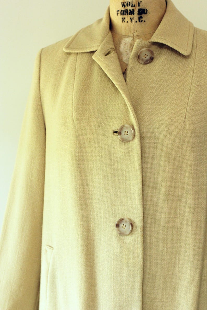 Puede incluir: Primer plano de un abrigo beige claro con cuello con solapas y tres botones grandes y redondos. El abrigo tiene una sutil textura de rayas verticales y un corte entallado. El abrigo se muestra en un maniqu&iacute; vintage.